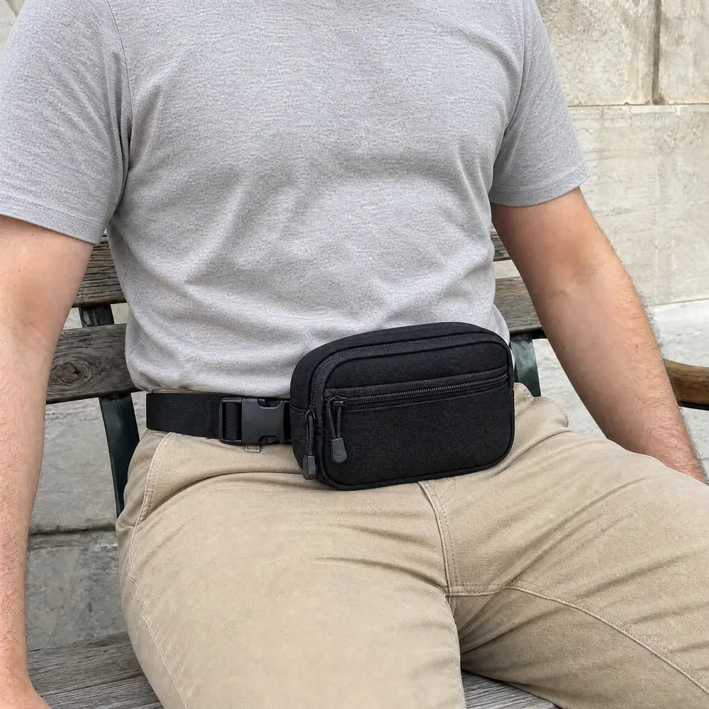 Adult seated on an outdoor bench wearing the closed black CloseCarry waist pouch with a grey T-shirt tucked into beige trousers.