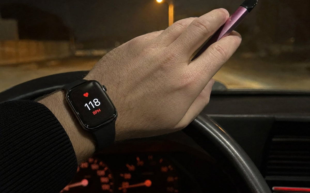 Smartwatch showing 118 BPM on a hand holding a vape on a steering wheel