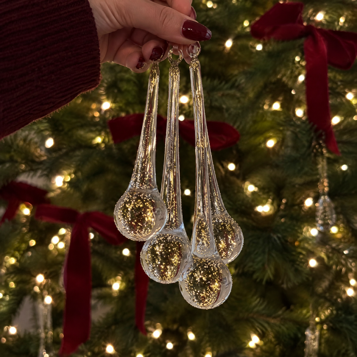 Hand holding four long clear glass drops in front of a lit tree