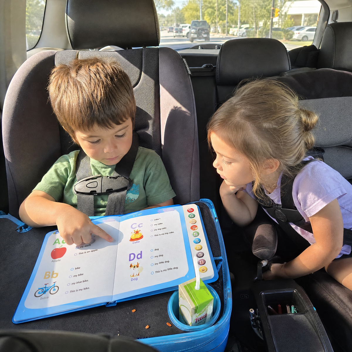 A boy reading in his car seat on a travel tray while his sister listens