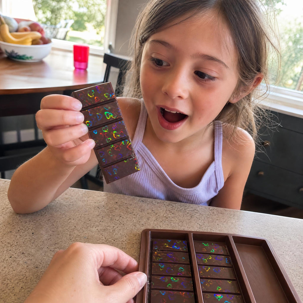 A girl gasps at a HoloPop chocolate bar shimmering with rainbow hearts while her mom holds the mold
