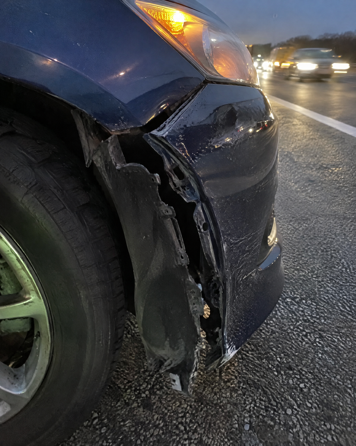 A torn bumper corner and hanging wheel-well liner on a highway shoulder at dusk
