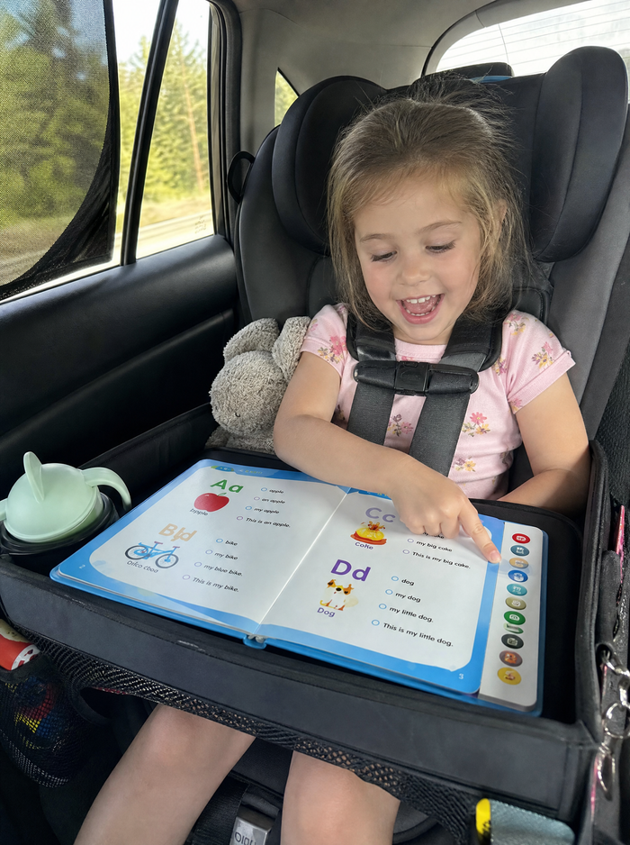 A girl in a car seat reading on a travel tray
