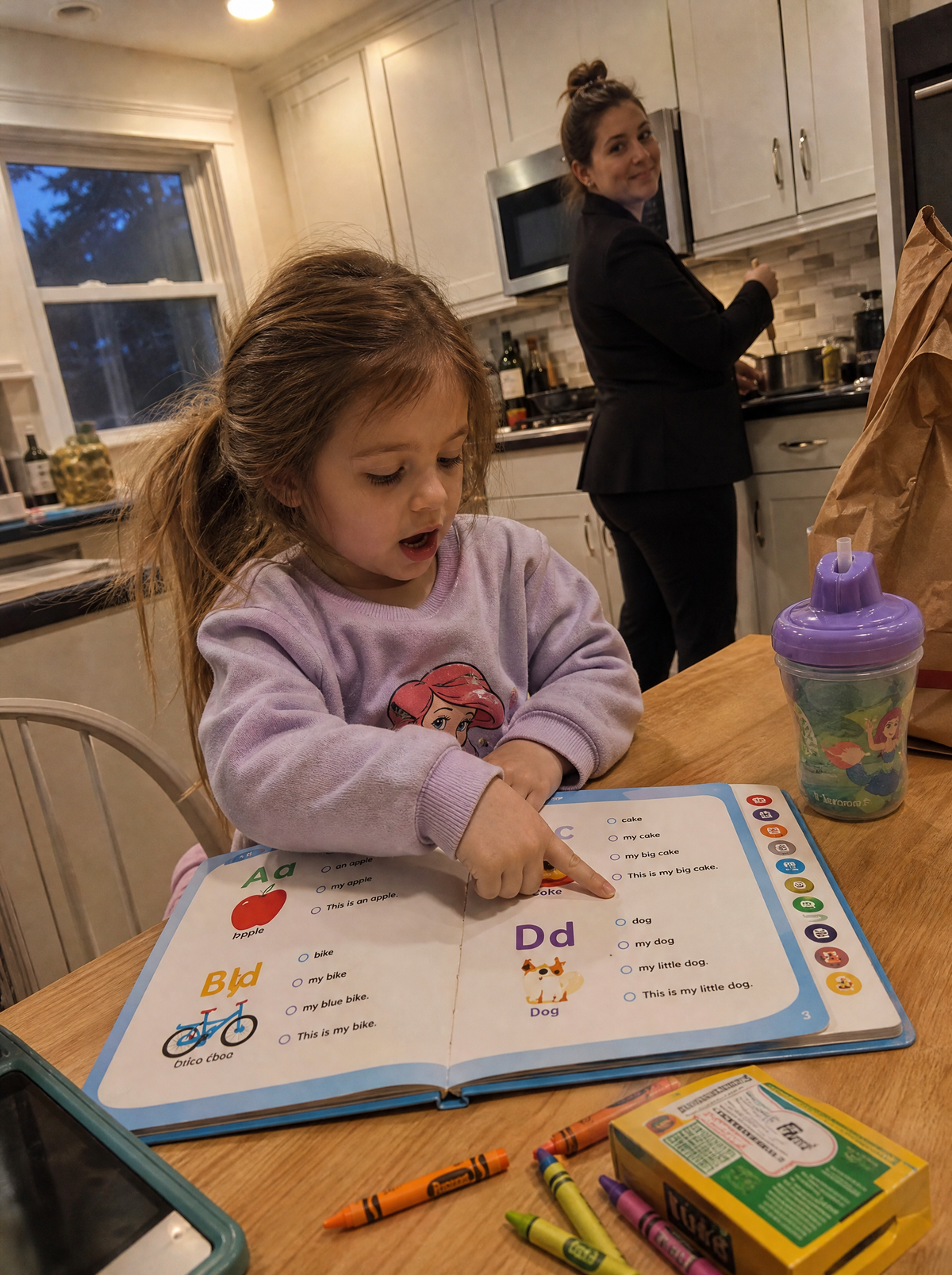 A girl reading with the talking book at the kitchen table while her mom cooks