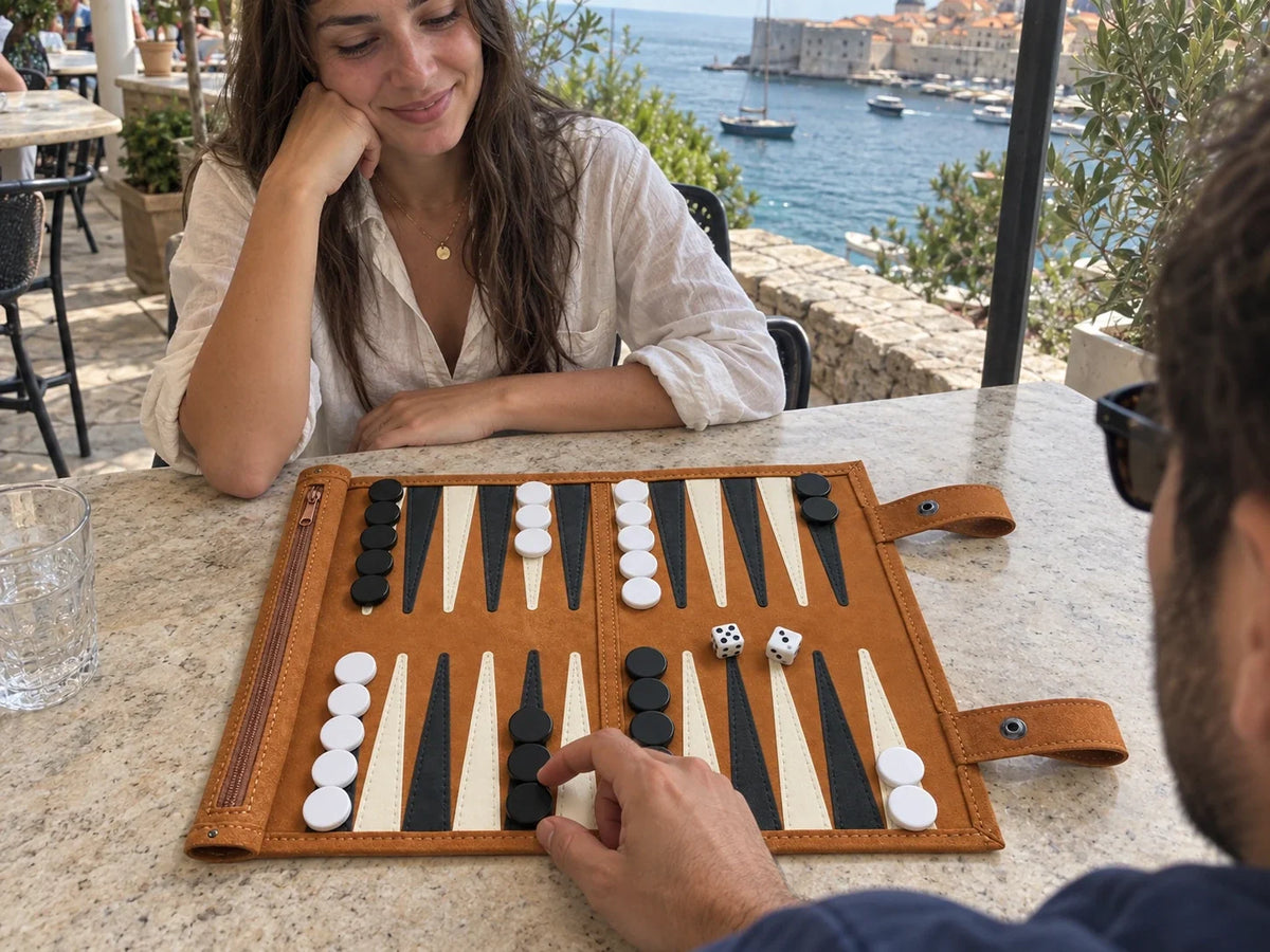 Two travelers seated opposite each other playing Ceyla Roam backgammon at a seaside cafe, with one player considering a move