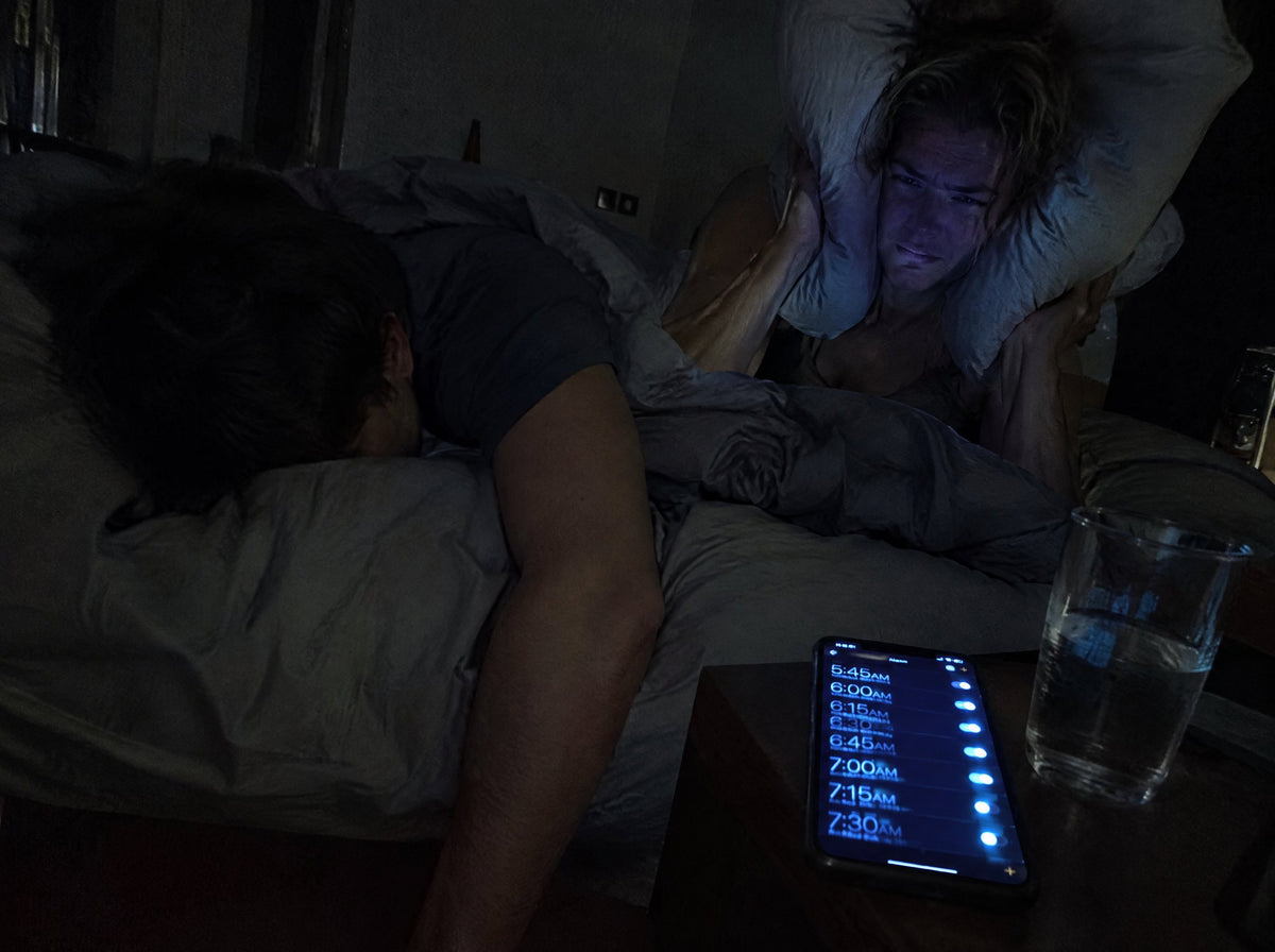 A man still asleep while his partner covers her ears as his phone alarms keep going off