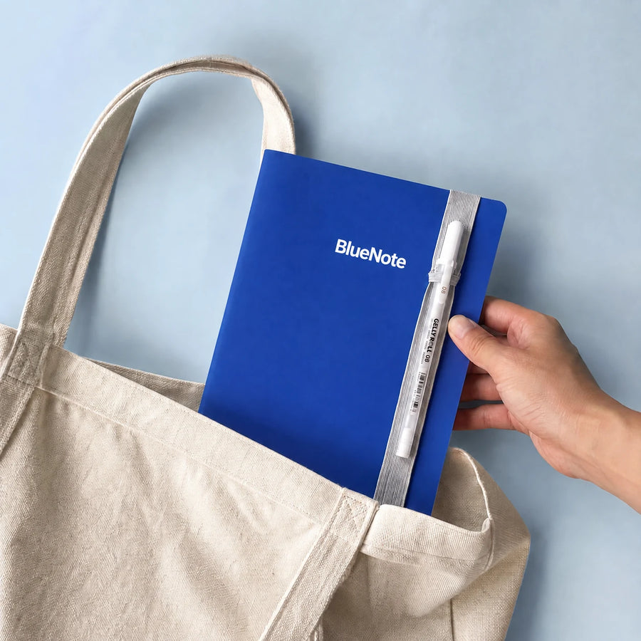BlueNote notebook with pen and strap being placed into a canvas tote; tote not included.
